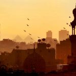The skyline of Cairo at sunset with birds over domes and satellite dishes with distant pyramids at Giza